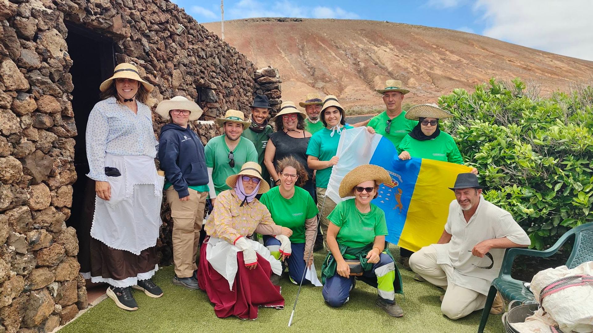 Equipo numeroso junto a construcción tradicional de piedra con montaña volcánica de tonos rojizos al fondo